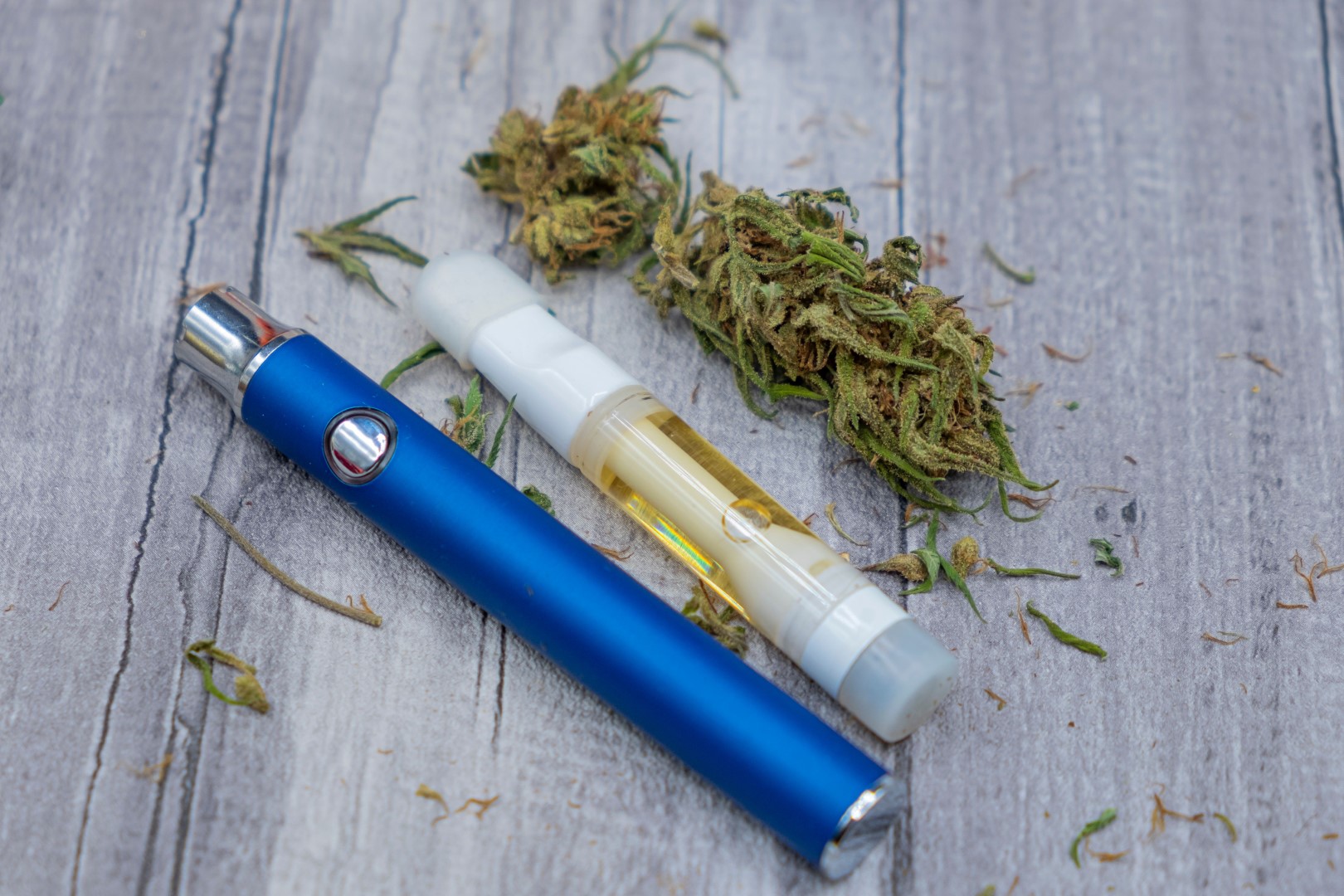 a blue vape 510 battery next to a pre filled cannabis concentrate cartridge and marijuana buds on a wooden background